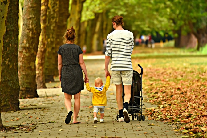 Ein Mann und eine Frau gehen, mit einem kleinen Kind an der Hand, spazieren. Der Mann schiebt einen Kinderwagen. Auf dem Boden liegt Herbstlaub.