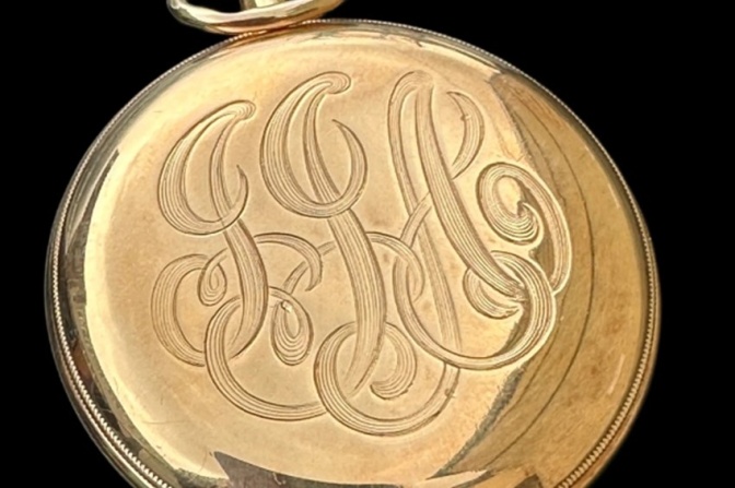 Eine goldene Taschenuhr mit geschlossenem Deckel. Darauf ist ein verschlungenes Muster aus drei ineinander verschlungenen J's zu sehen.