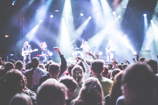 Eine große Menge an Zuschauer*innen vor einer Festivalbühne. Manche von ihnen heben die Arme in die Luft. Die Bühne ist von verschiedenen Scheinwerfern bläulich beleuchtet.