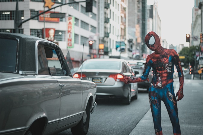 Ein Mann im Spiderman Kostüm steht an der Straße und streckt den Daumen nach oben, um von einem Auto mitgenommen zu werden.