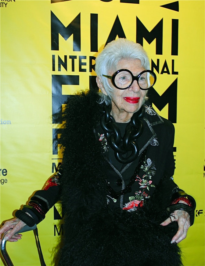 Iris Apfel in schwarzer Kleidung mit Federboa und großer schwarzer Brille. Sie steht lächelnd vor einem leuchtend gelben Filmplakat.