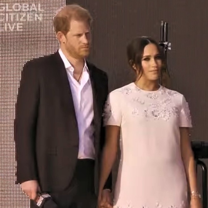Prinz Harry und Herzogin Meghan stehen händchenhaltend auf einer Bühne. Sie tragen festliche Kleidung, Harry hat ein Mikro in der Hand.