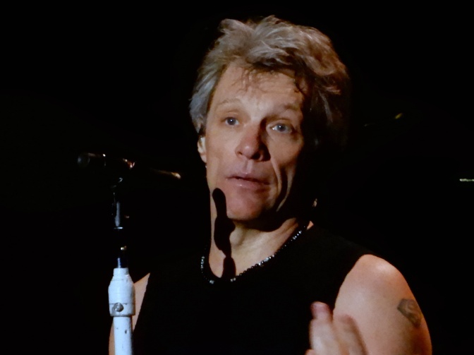 Jon Bon Jovi in schwarzem T-Shirt auf der Bühne am Mikrofon.