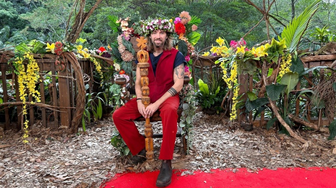Gil Ofarim trägt einen Blumenkranz und hat eine großen Holzstab in der Hand. Er sitzt auf einem Thron und lächelt.