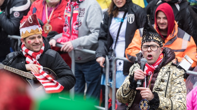 Menschen mit Narrenkappen und rot-weißen Schals am Straßenrand. Ein Mann mit Narrenkappe spricht in ein Mikrofon.