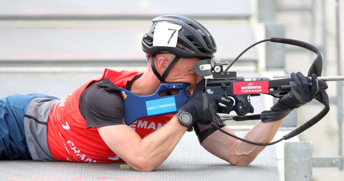 Ein Mann mit Helm in rotem Trikot liegt auf dem Boden und schießt auf ein Ziel.