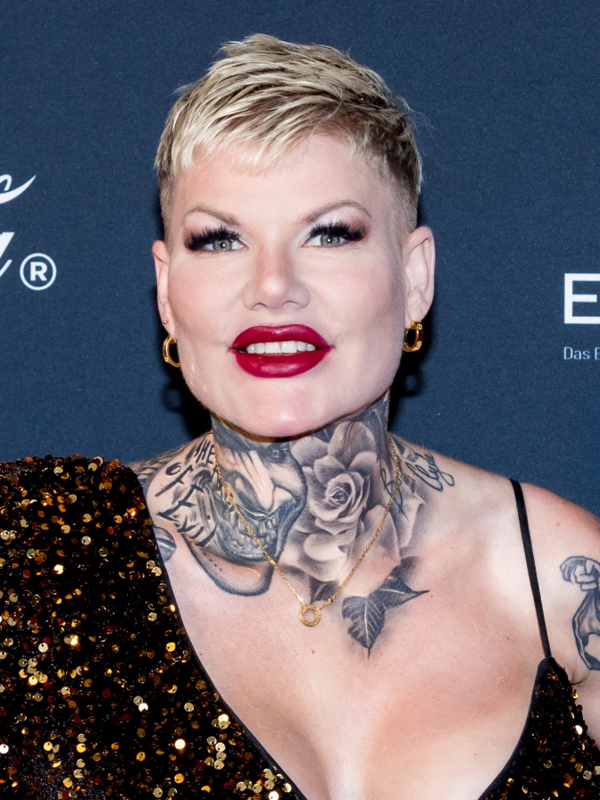 Melanie Müller mit einem großen Halstattoo und kurzen blondierten Haaren. Sie steht vor einer Logowand und trägt ein asymmetrisches schwarzes Kleid mit Glitzer.
