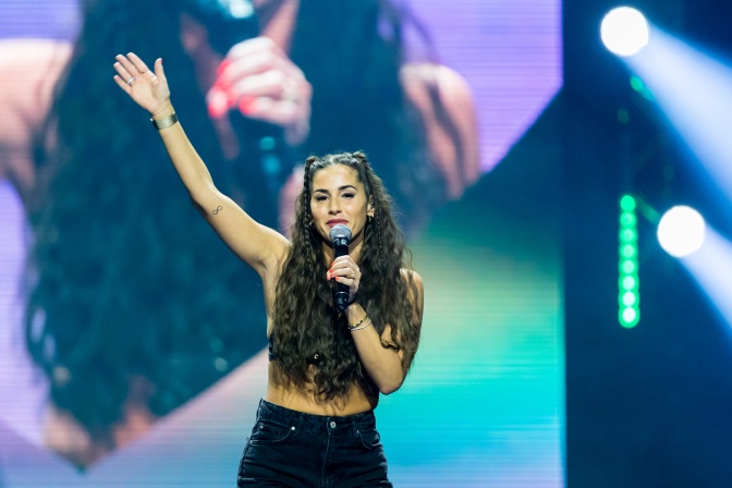 Sarah Engels steht auf der Bühne vor einer riesigen Leinwand. In einer Hand hält sie ein Mikro, mit der anderen Hand winkt sie ins Publikum.