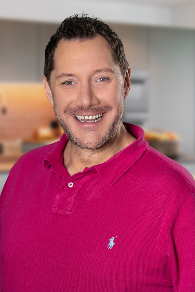Mirko Reeh mit Schnurrbart und Dreitagebart in einem pinkfarbenen Poloshirt. Er lächelt in die Kamera.