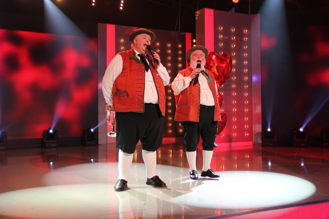 2 dicke Männer in rotweißer Tracht auf der Bühne. Sie singen in Mikrofone.