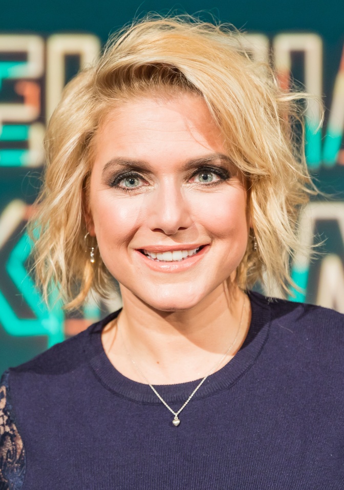 Jeanette Biedermann mit kinnlangen, leicht gewellten blonden Haaren. Sie steht lächelnd vor einer Logowand.