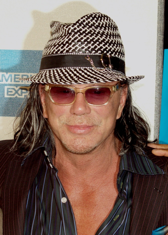 Mickey Rourke mit langen Haaren, Hut und Sonnenbrille. Er hat einen Dreitagebart und lächelt leicht in die Kamera.