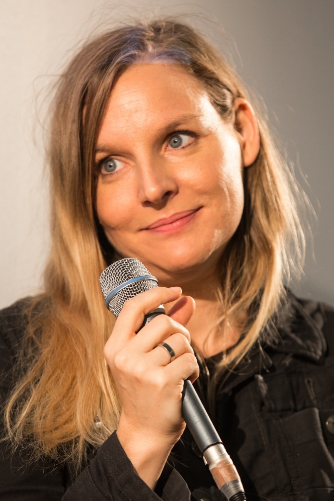 Judith Holofernes hat ein Mikrofon in der Hand, hat den Kopf leicht schief gelegt und lächelt leicht. Sie schaut seitlich an der Kamera vorbei.