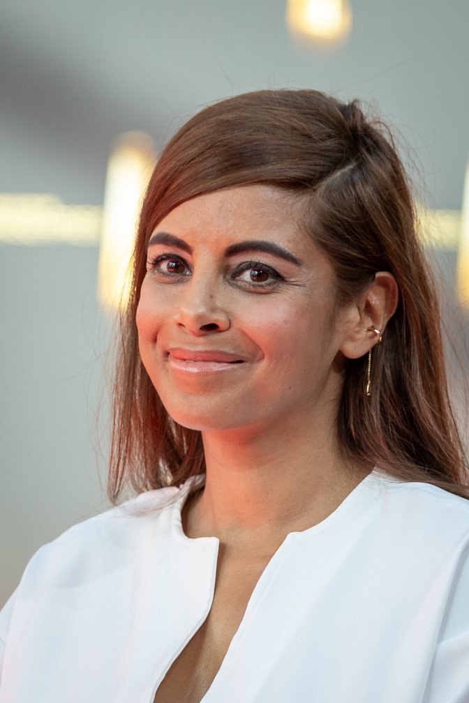 Collien Fernandes mit dunklen, langen Haaren in einer weißen Jacke. Sie steht lächelnd vor einer Logowand.