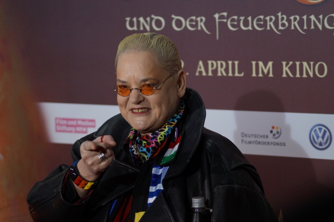 Hella von Sinnen mit einer orangefarbenen Sonnenbrille und nach hinten frisierten hellblonden Haaren. Sie steht vor einem Kinoplakat und zeigt lächelnd auf den Fotografen.