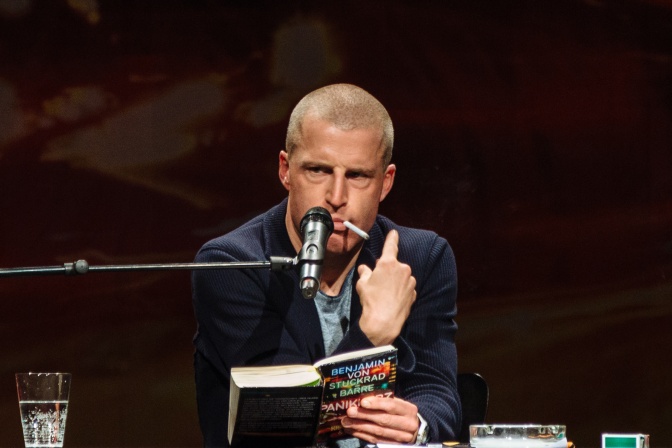 Benjamin von Stuckrad-Barre sitzt vor einem Mikro, hat eine nicht angezündete Zigarette im Mund und liest aus einem Buch.