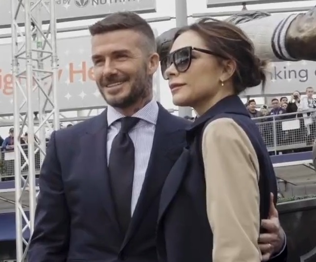 David und Victoria Beckham stehen nebeneinander. Er hat den Arm auf ihre Hüfte gelegt. Sie trägt eine große schwarze Sonnenbrille.