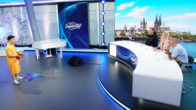 Eine Person steht vor dem riesigen Jury-Tisch der Sendung DSDS, die 3 Jury-Mitglieder sitzen dahinter.