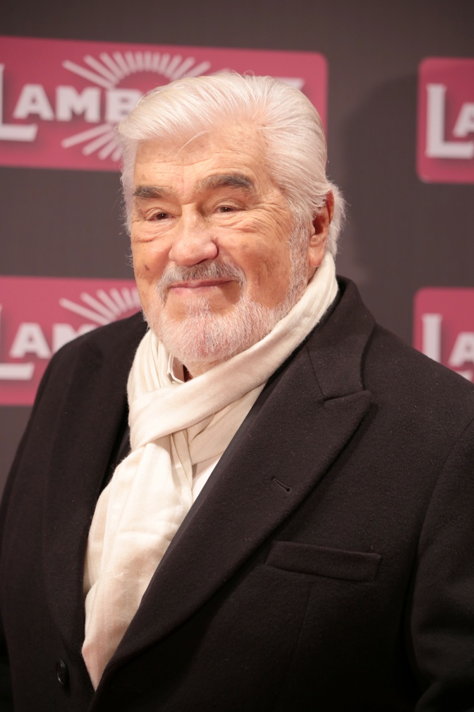 Mario Adorf mit weißem Schal und schwarzem Mantel vor einer Logowand. Er hat weiße Haare und trägt einen weißen Vollbart.
