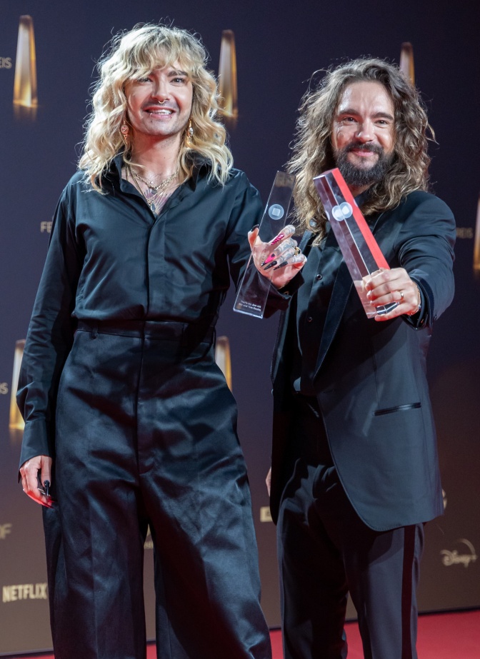 Bill und Tom Kaulitz stehen in schwarzer Kleidung nebeneinander auf der Bühne und halten eine Figur aus Plexiglas in der Hand.