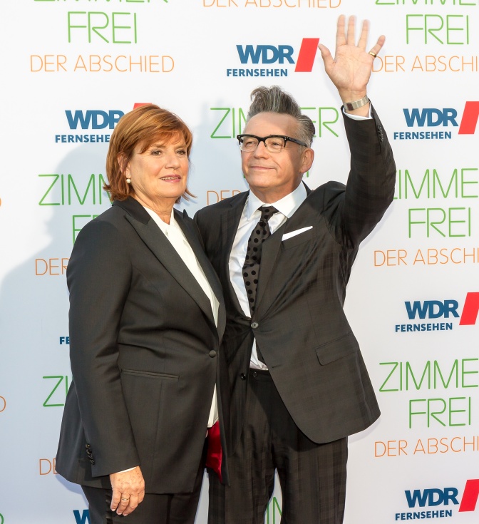 Christine Westermann und Götz Alsmann in dunklen Anzügen vor einer Logowand des WDR. Götz Alsmann winkt.