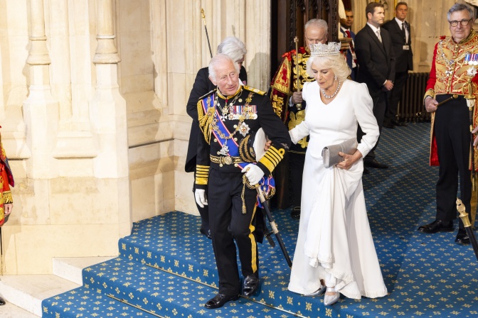 Charles und Camilla verlassen gemeinsam ein Gebäude. Sie trägt eine Krone, er zahlreiche Orden.