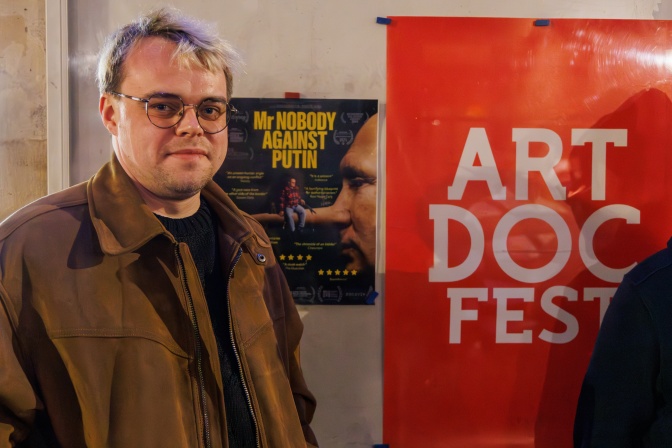 Pavel Talankin mit blondierten kurzen Haaren und Brille. Er steht neben einem Banner des Art Doc Fests.