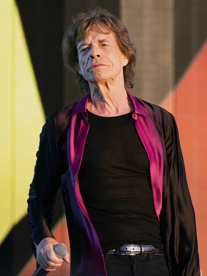 Mick Jagger mit einem schwarzen Blazer mit pinkfarbenen Revers. Er steht sehr aufrecht und schaut provozierend in die Kamera.