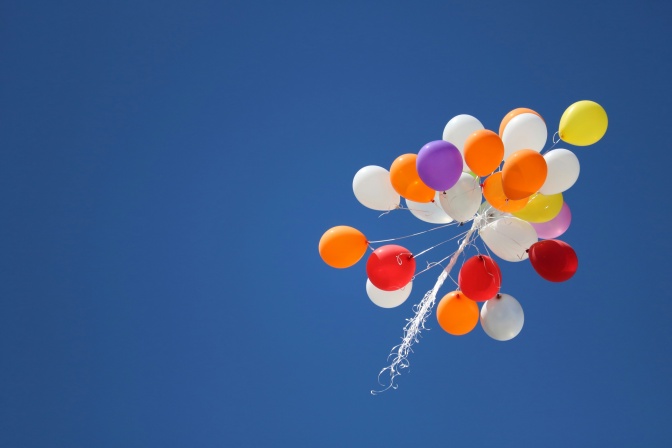 Bunte Luftballons sind zu einem Strauß zusammengebunden und fliegen in einem leuchtend blauen, wolkenlosen Himmel.