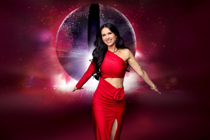 Vanessa Borck in einem engen roten Kleid mit Cutouts. Sie breitet beide Arme aus und lächelt in die Kamera.