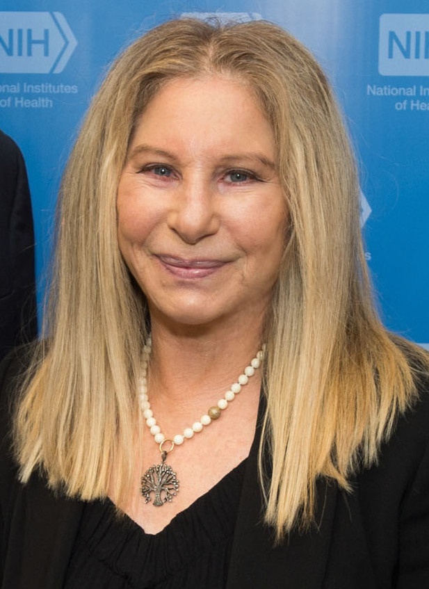 Barbra Streisand mit glatten blonden Haaren in einem schwarzen Kleid vor einer Logowand.