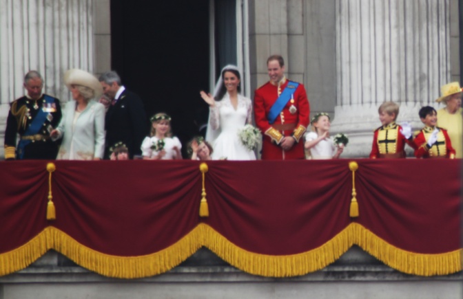 Das Paar steht auf einem Balkon und winkt. Um sie herum stehen die Mitglieder der britischen Königsfamilie.