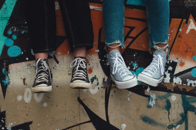 Vier Beine und Füße baumeln vor einer mit Graffiti besprayten Mauer. Die beiden Personen tragen Jeans und Schuhe von Converse.
