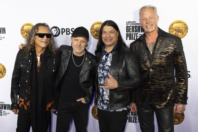 Die 4 Mitglieder der Band Metallica in schwarzer Kleidung vor einer Logowand.