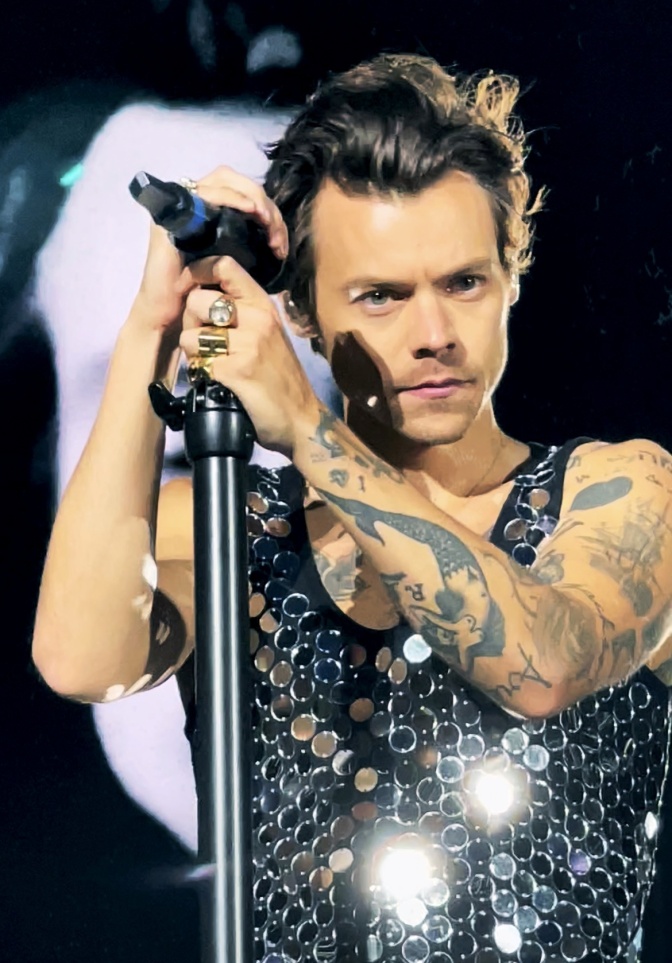 Harry Styles in einem Shirt mit großen silbernen Pailletten. Er steht an einem Standmikro auf der Bühne.