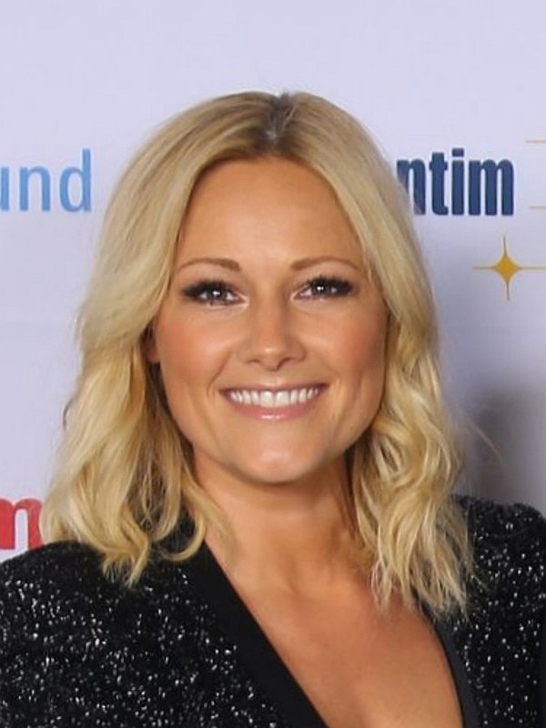 Helene Fischer mit schulterlangen blonden gewellten Haaren in einem schwarzen Oberteil. Sie steht lächelnd vor einer Logowand.