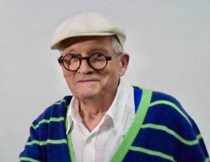 David Hockney als alter Mann. Er trägt eine weiße Kappe, Brille und einen gestreiften Pullover.