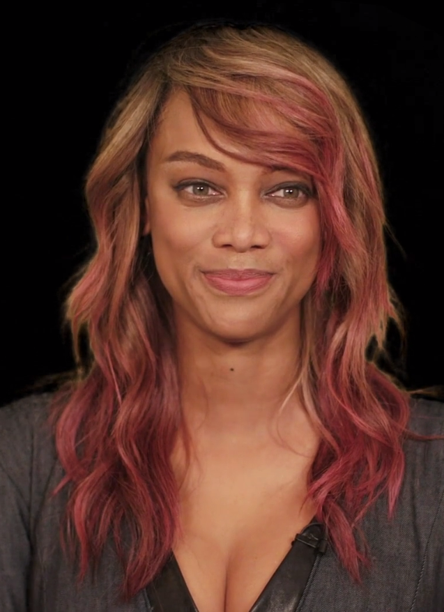 Tyra Banks mit langen, gewellten rotbraunen Haaren. Sie lächelt und schaut zur Seite.
