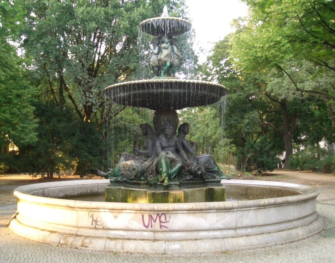 Ein runder Brunnen, außen aus hellem Marmor, innen aus grünlich angelaufenem Kupfer. In der Mitte sitzen 4 große Figuren aus hellem Stein.