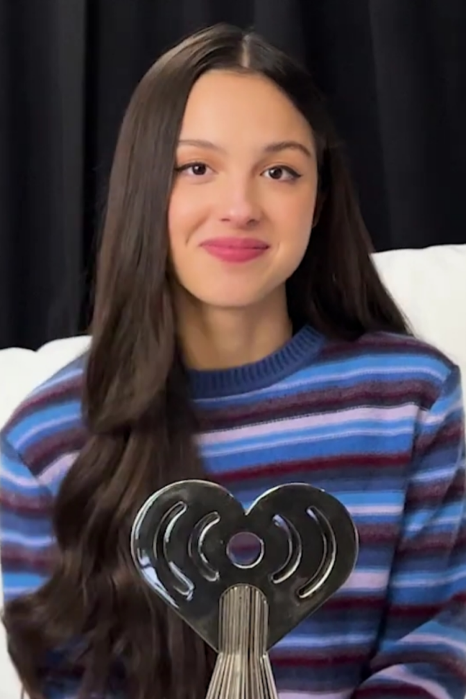 Olivia Rodrigo in einem gestreiften Pullover mit langen, glatten schwarzen Haaren. Vor ihr steht eine silberne, herzförmige Statue.