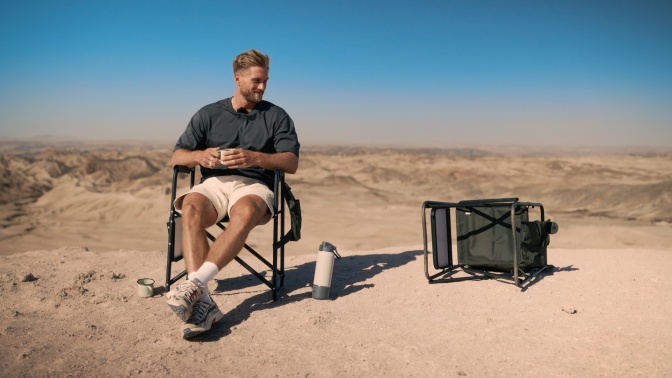 Ein blonder, bärtiger Mann sitzt im Freien auf einem Campingstuhl. Neben ihm liegt ein zweiter Campingstuhl, der vom Wind umgeweht wurde.