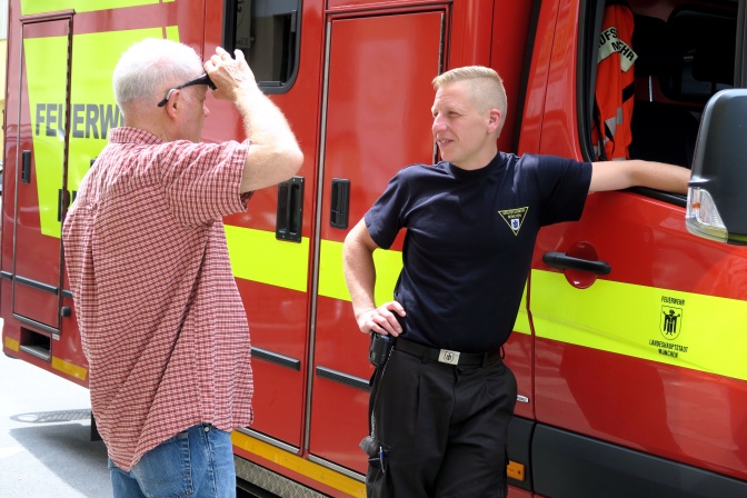 Ein Mann in Uniform und ein Mann in rot-weiß-kariertem Hemd stehen vor einem roten Feuerwehrauto.