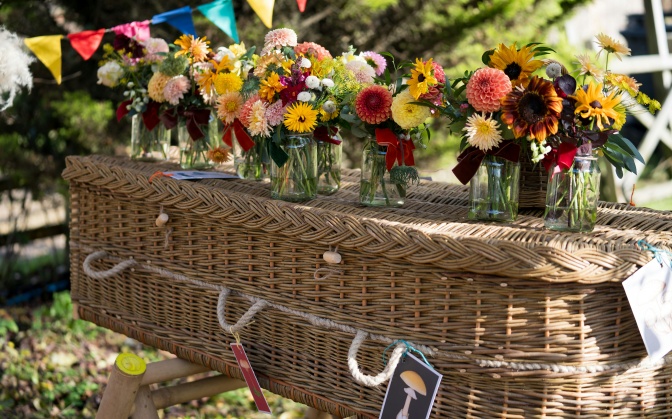 Ein Sarg aus geflochtenem Kord. Darauf stehen flache Glasvasen mit Blumen.