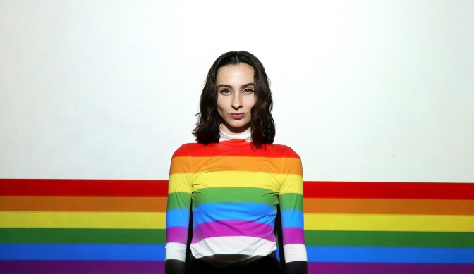 Eine Frau steht vor einer weißen Wand und wird von Streifen in Regenbogenfarben angeleuchtet. Sie sehen aus wie Streifen auf ihrem Pulllover, ziehen sich aber über die ganze Wand.