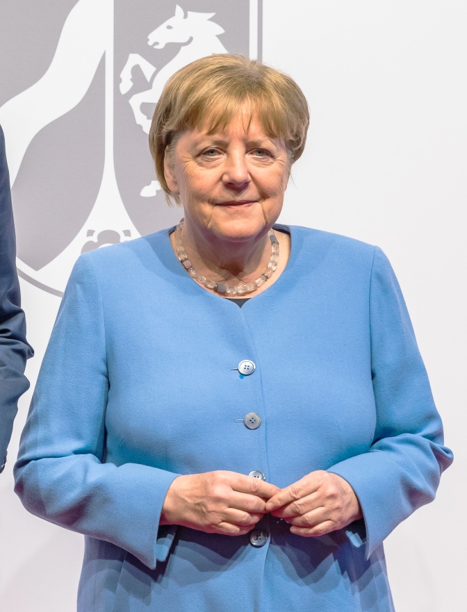 Angela Merkel in einem hellblauen Blazer. Sie steht vor dem Wappen von Nordrhein Westfalen und schaut in die Kamera.