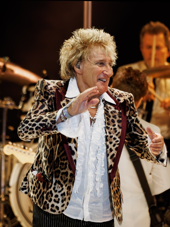 Rod Stewart in einem Sakko mit Leopardenmuster und in einem weißen Hemd mit Rüschen. Er steht auf der Bühne.