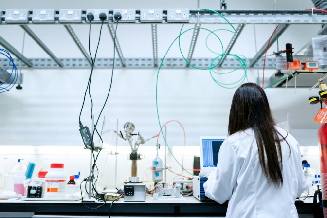 Eine weibliche Wissenschaftlerin in einem Elektrolabor. Sie trägt einen weißen Kittel und ist von hinten zu sehen.