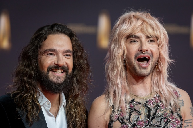 Tom und Bill Kaulitz vor einer Logowand. Beide lächeln in die Kamera.