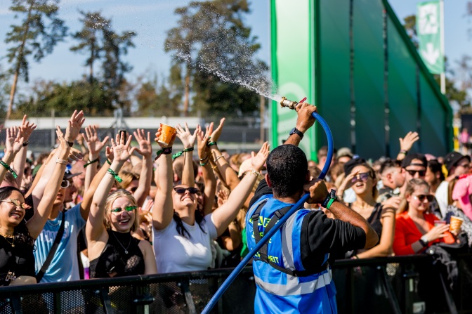 Festivalbesucher*innen in sommerlicher Kleidung. Ein Mann in blauer Weste bespritzt sie zur Abkühlung mit einem Wasserschlauch.