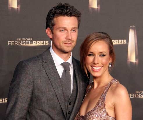 Annemarie und Wayne Carpendale in festlicher Kleidung vor einer Logowand. Sie stehen eng beieinander und lächeln in die Kamera.
